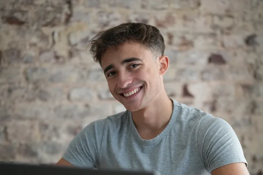 Bastian Holthausen lacht im grauen T-Shirt am Laptop vor einer Backsteinwand