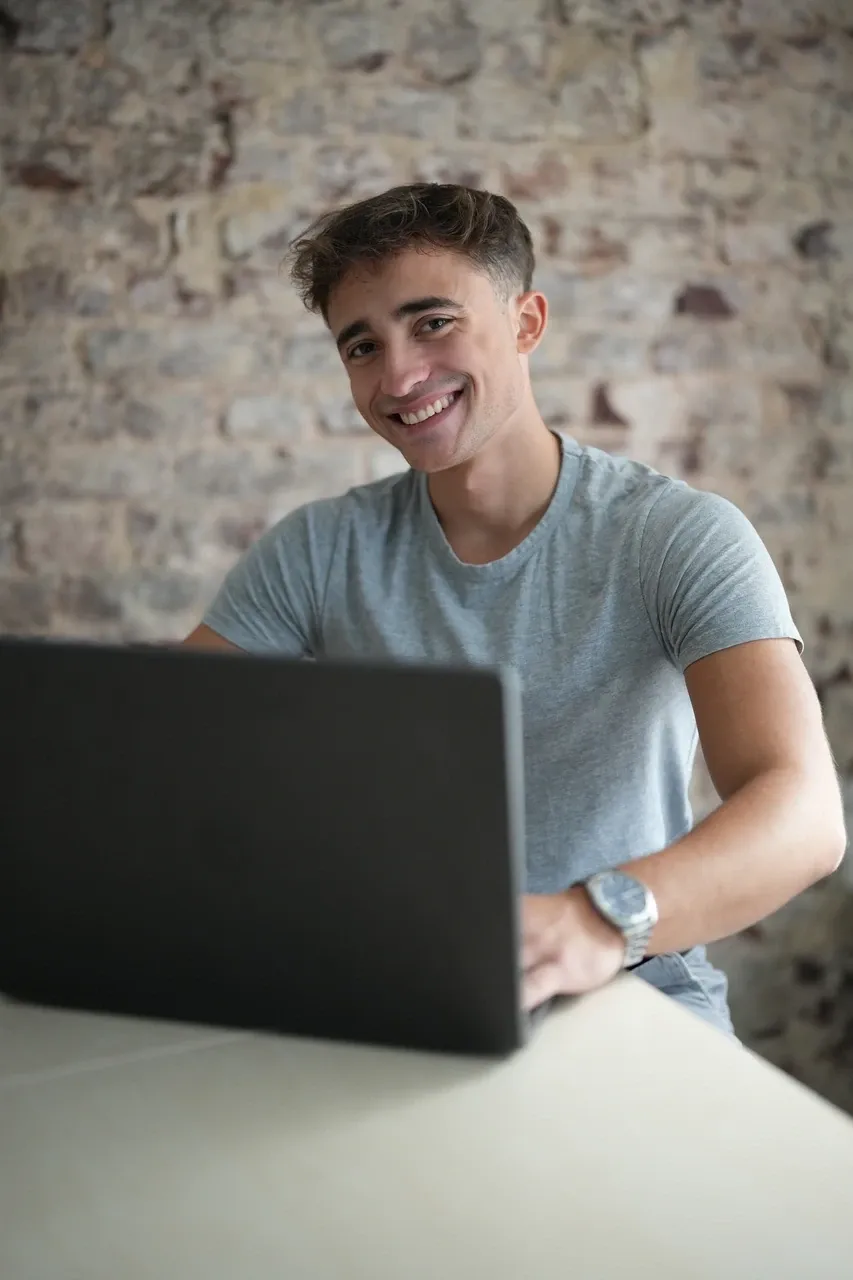 Bastian Holthausen, Inhaber von Noxoros, sitzt lächelnd am Laptop vor einer Backsteinwand