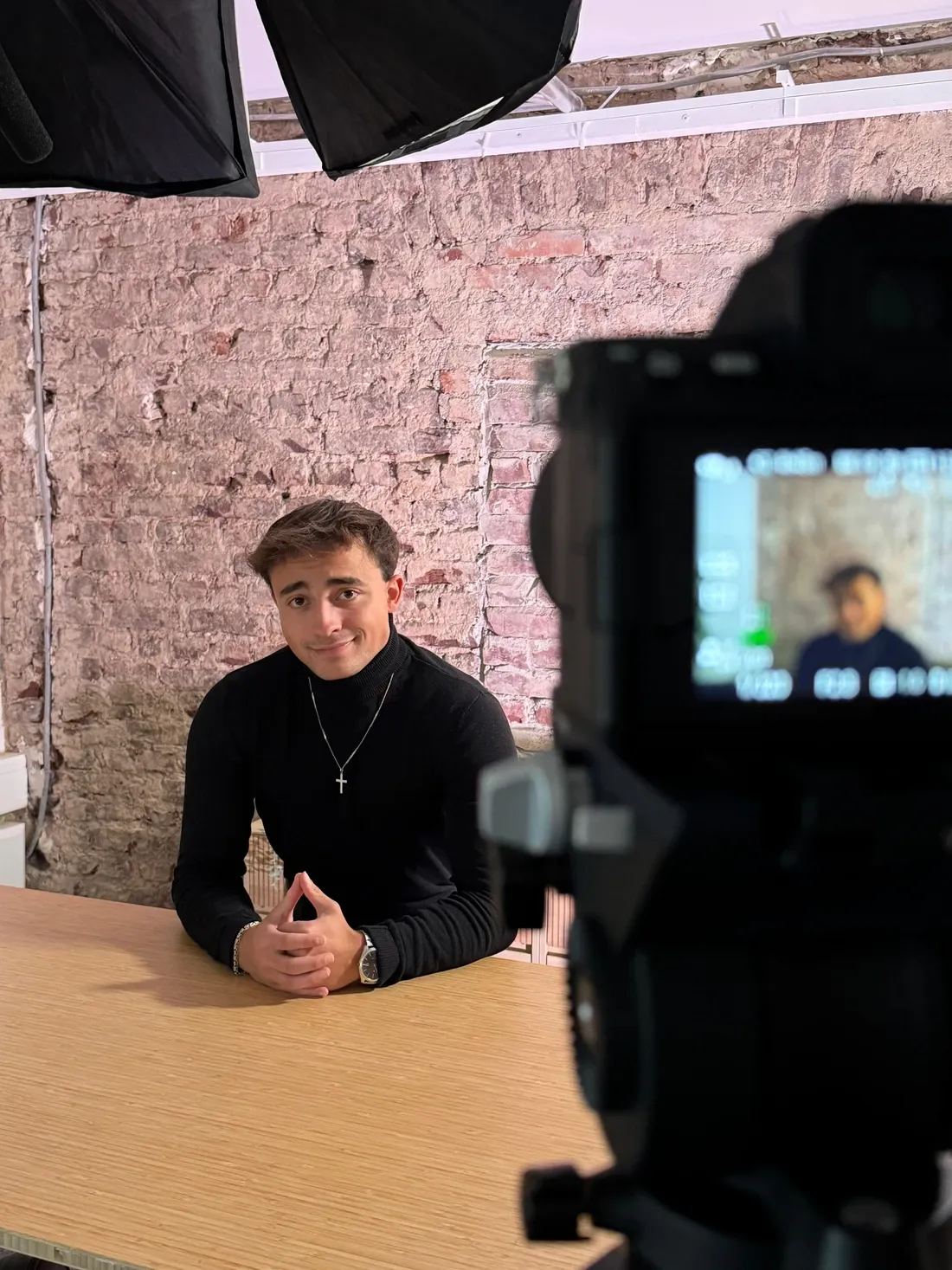 Bastian Holthausen im schwarzen Rollkragenpullover am Holztisch vor einer Backsteinwand, davor eine Kamera beim Videodreh