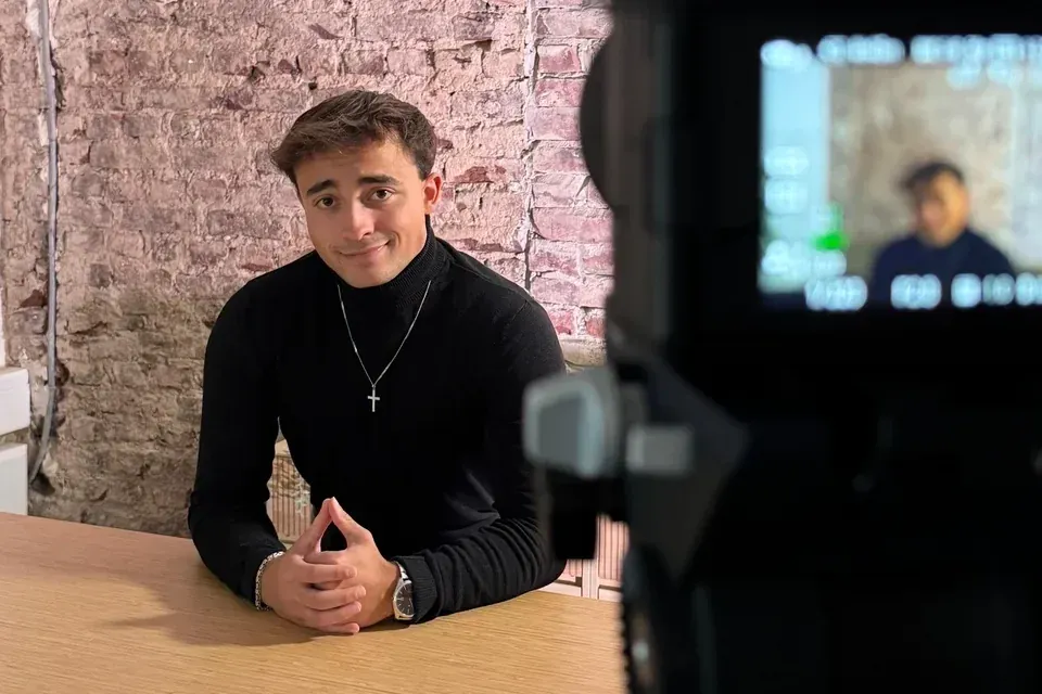 Bastian Holthausen im schwarzen Rollkragenpullover am Holztisch vor einer Backsteinwand, daneben eine Kamera beim Videodreh